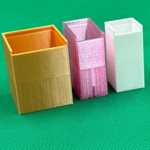 May include: Three open-top rectangular containers arranged in a row. The first is a wide gold-colored box, the second is a translucent pink box, and the third is a narrow white box. Each container features a smooth, layered 3D-printed texture.