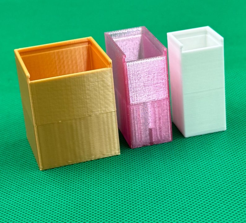 May include: Three open-top rectangular containers arranged in a row. The first is a wide gold-colored box, the second is a translucent pink box, and the third is a narrow white box. Each container features a smooth, layered 3D-printed texture.
