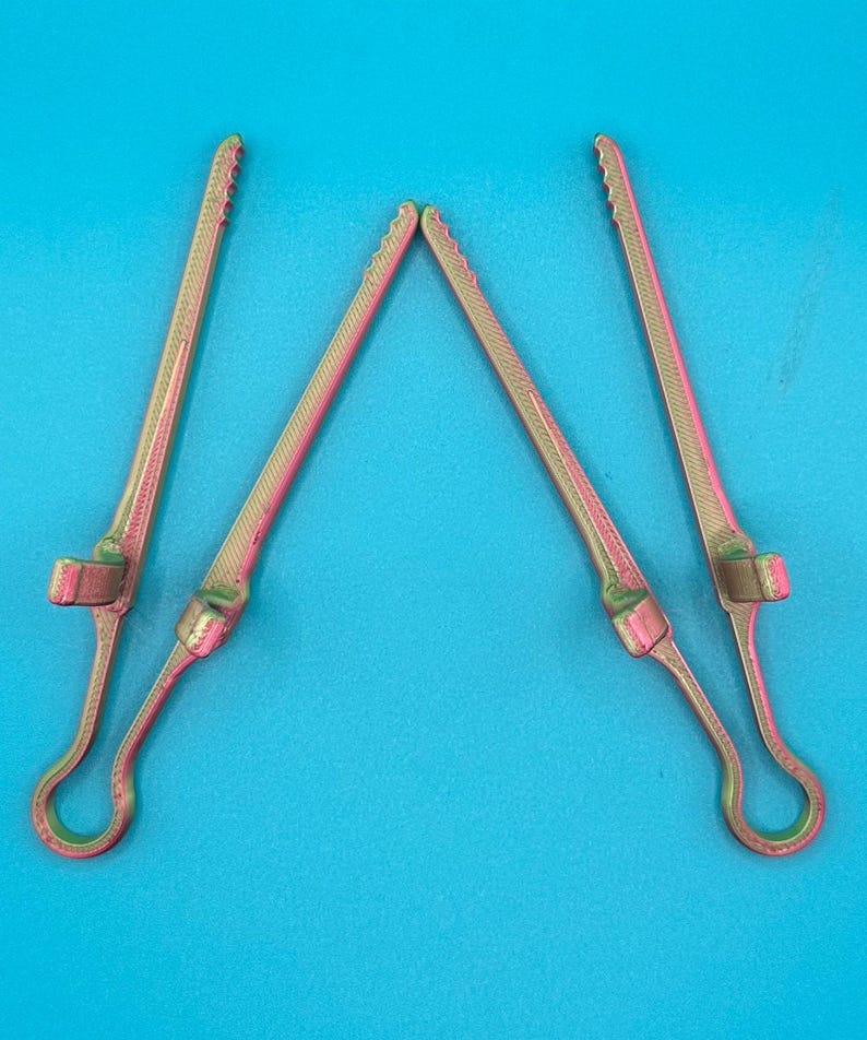 May include: Four long, slender, metallic gold and pink iridescent clips with a textured surface. Each clip features a circular loop at one end, a small perpendicular tab near the base, and a serrated edge along the top length of the arm.