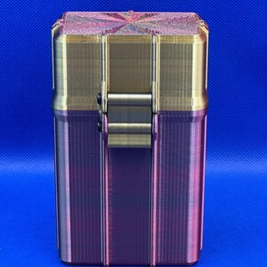 May include: A rectangular storage container with a hinged lid, featuring a metallic gold upper section and a gradient purple and dark pink lower section. The lid has a textured, starburst-like pattern on top, and a central latch secures the two halves.