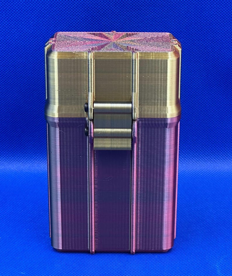 May include: A rectangular storage container with a hinged lid, featuring a metallic gold upper section and a gradient purple and dark pink lower section. The lid has a textured, starburst-like pattern on top, and a central latch secures the two halves.