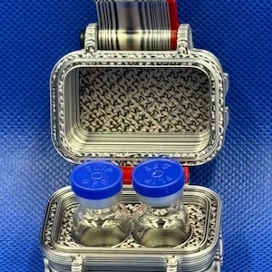 May include: A silver-colored, rectangular, 3D-printed storage case with a hinged lid, featuring a textured interior pattern. Inside, two small, clear glass vials with blue plastic caps labeled "FLIP OFF" are held in place.