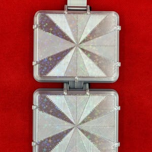 May include: A rectangular, two-part hinged object with a silver-toned frame. Each section features a holographic, star-patterned inlay with a radiating, sunburst-like design of alternating textured and smooth triangular segments.