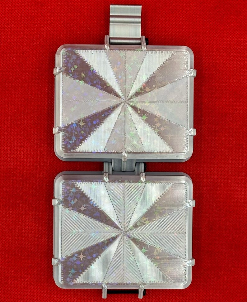 May include: A rectangular, two-part hinged object with a silver-toned frame. Each section features a holographic, star-patterned inlay with a radiating, sunburst-like design of alternating textured and smooth triangular segments.