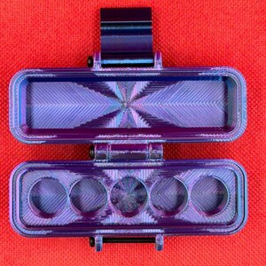 Puede incluir: Un estuche de almacenamiento rectangular con bisagras, impreso en 3D, con un acabado iridiscente met&aacute;lico en tonos morado y azul. La secci&oacute;n superior es un compartimento abierto y la inferior tiene cinco aberturas circulares en fila.