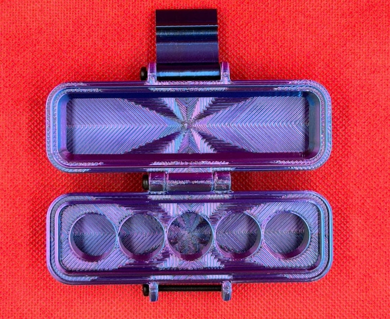 Puede incluir: Un estuche de almacenamiento rectangular con bisagras, impreso en 3D, con un acabado iridiscente met&aacute;lico en tonos morado y azul. La secci&oacute;n superior es un compartimento abierto y la inferior tiene cinco aberturas circulares en fila.