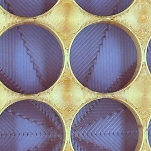 May include: A close-up of a gold-colored metal grid with circular openings. The openings are filled with a purple material that has a textured, layered appearance.