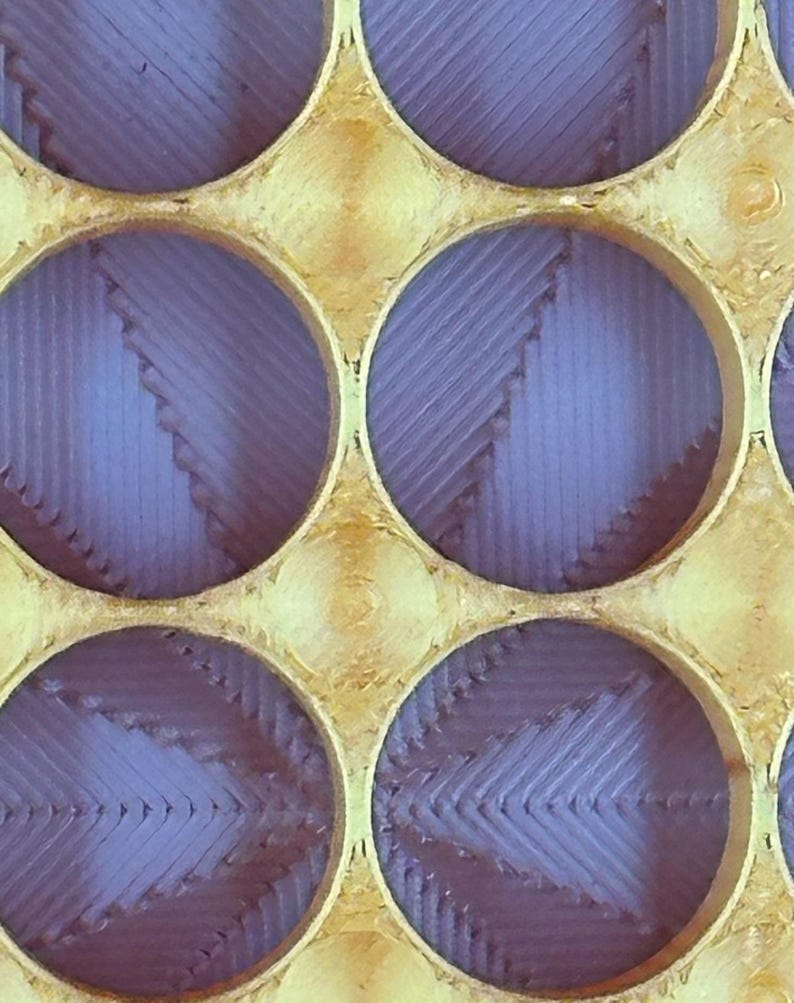 May include: A close-up of a gold-colored metal grid with circular openings. The openings are filled with a purple material that has a textured, layered appearance.