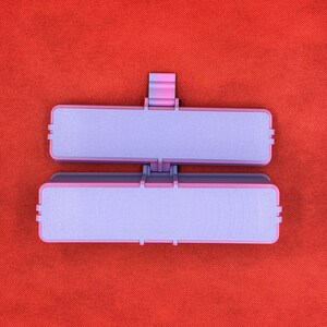 May include: A rectangular, two-part hinged container featuring a light purple interior and a darker purple exterior frame. The object has a small, centered latch mechanism on the top edge, designed to securely close the two halves together.