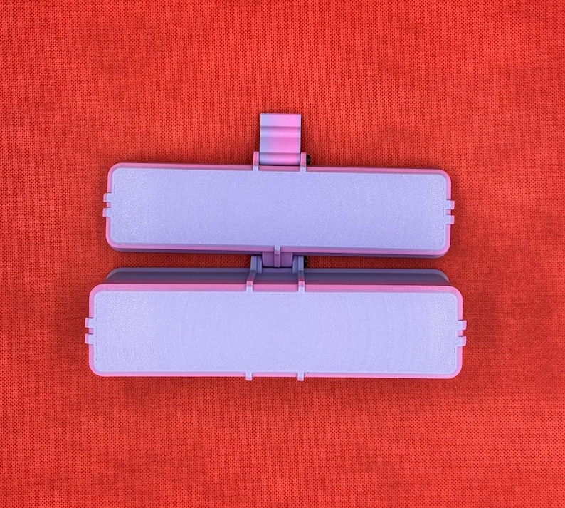 May include: A rectangular, two-part hinged container featuring a light purple interior and a darker purple exterior frame. The object has a small, centered latch mechanism on the top edge, designed to securely close the two halves together.