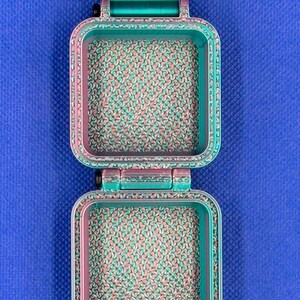 May include: A square, hinged container featuring a gradient color scheme that transitions from teal to magenta. The interior surfaces and the outer frame display a textured, repeating pattern. A solid teal rectangular tab extends from the top.