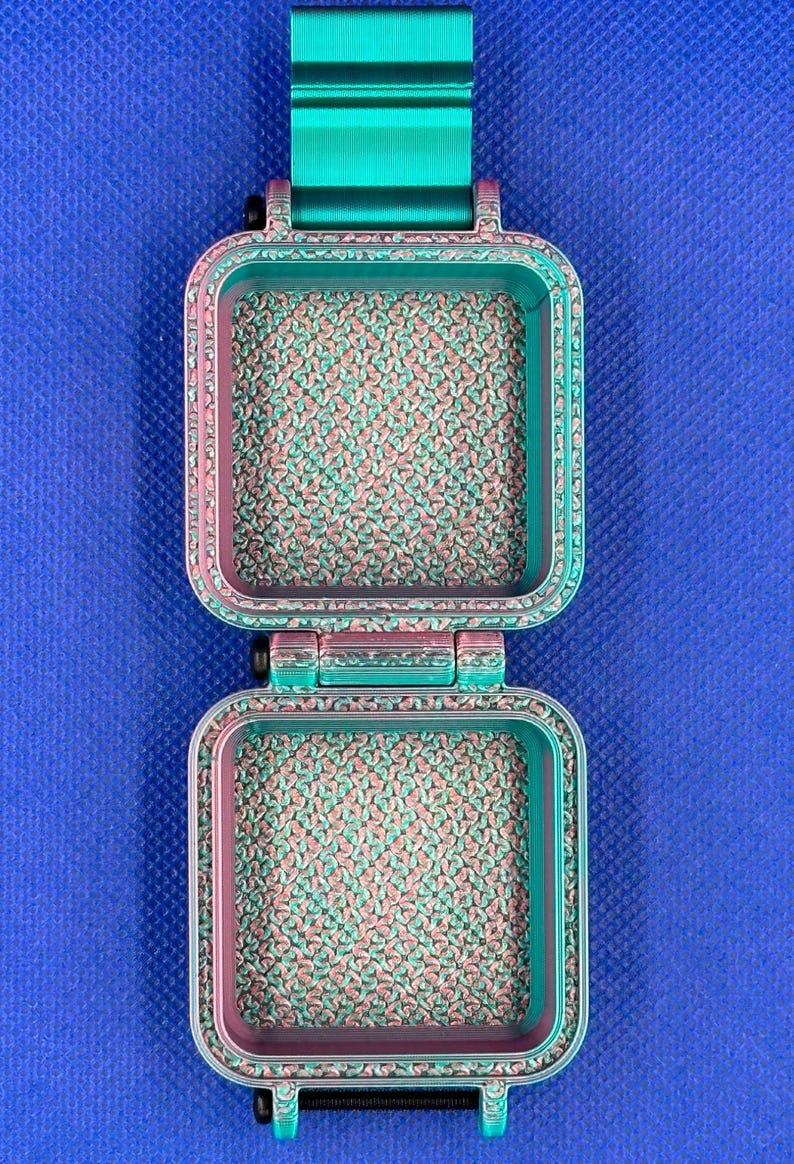 May include: A square, hinged container featuring a gradient color scheme that transitions from teal to magenta. The interior surfaces and the outer frame display a textured, repeating pattern. A solid teal rectangular tab extends from the top.