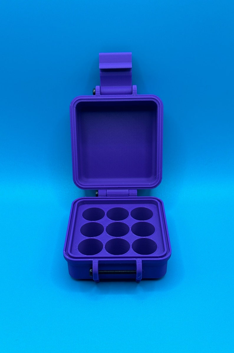 May include: A purple plastic storage case with a hinged lid, featuring an open interior compartment with a grid of nine circular holes arranged in a three-by-three pattern. The case includes a latch mechanism and a visible black metal hinge pin.