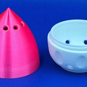 May include: A pink and white 3D printed egg-shaped container with a pointed top and a round bottom. The top has several small holes. The bottom has a textured surface with small bumps.