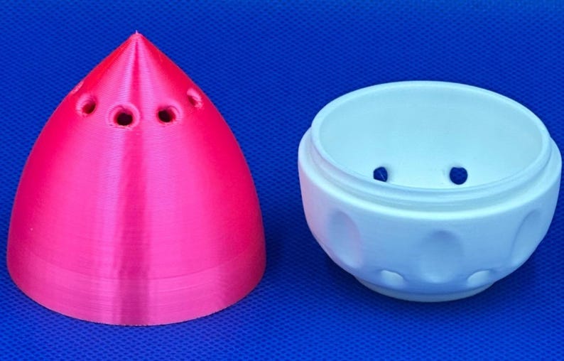 May include: A pink and white 3D printed egg-shaped container with a pointed top and a round bottom. The top has several small holes. The bottom has a textured surface with small bumps.