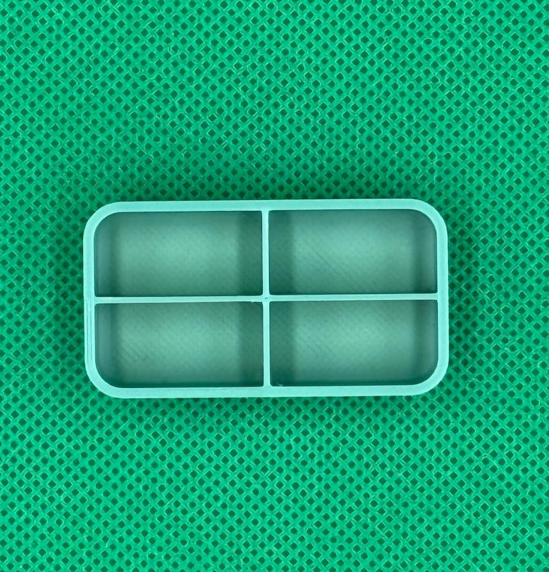 May include: A light teal rectangular silicone mold with four equally sized compartments, perfect for crafting. The mold has rounded corners and is set against a textured green background. Ideal for creating small, uniform shapes.