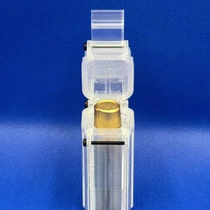 Puede incluir: Un estuche de almacenamiento rectangular y transparente de pl&aacute;stico impreso en 3D con tapa abatible, que contiene un objeto met&aacute;lico cil&iacute;ndrico con una parte superior dorada. El estuche est&aacute; asegurado con peque&ntilde;os tornillos negros.