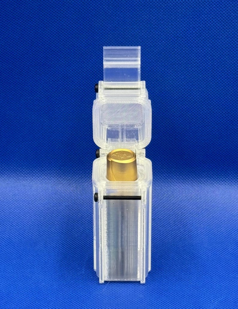 Puede incluir: Un estuche de almacenamiento rectangular y transparente de pl&aacute;stico impreso en 3D con tapa abatible, que contiene un objeto met&aacute;lico cil&iacute;ndrico con una parte superior dorada. El estuche est&aacute; asegurado con peque&ntilde;os tornillos negros.