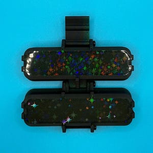 Puede incluir: Un accesorio negro de doble bisagra con dos segmentos rectangulares de esquinas redondeadas. Cada segmento tiene una superficie oscura con un patr&oacute;n de estrellas hologr&aacute;ficas que refleja varios colores. Incluye un clip central vertical.