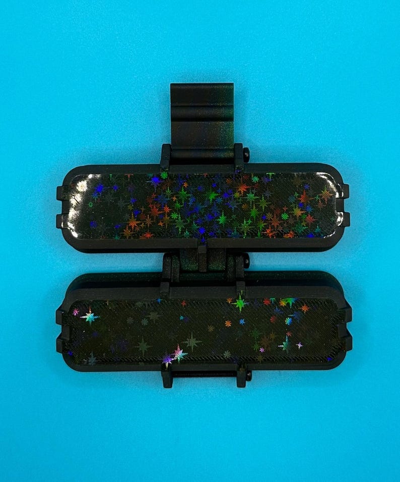 Puede incluir: Un accesorio negro de doble bisagra con dos segmentos rectangulares de esquinas redondeadas. Cada segmento tiene una superficie oscura con un patr&oacute;n de estrellas hologr&aacute;ficas que refleja varios colores. Incluye un clip central vertical.