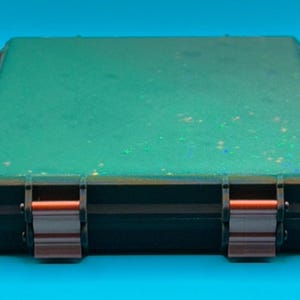 May include: A rectangular, flat, black storage case featuring a vibrant green top surface embedded with fine, shimmering glitter particles. The case is secured by two metallic, copper-coloured latches positioned on the front edge.