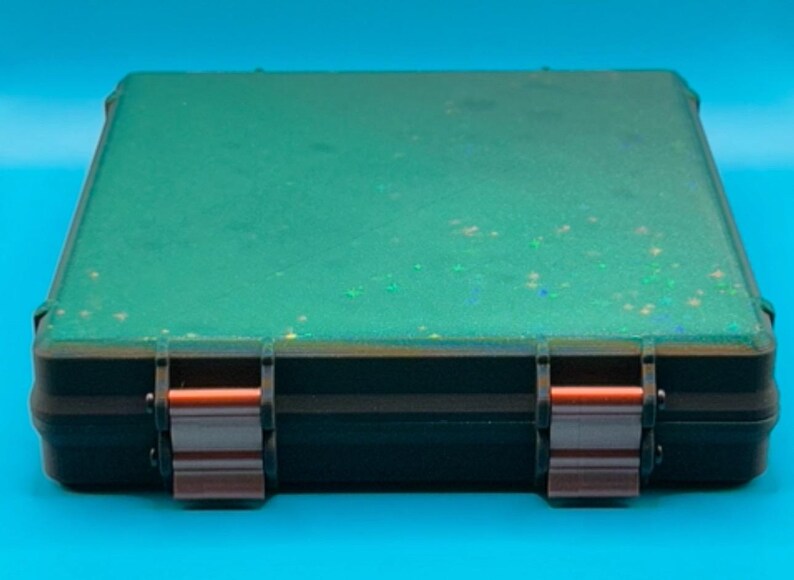 May include: A rectangular, flat, black storage case featuring a vibrant green top surface embedded with fine, shimmering glitter particles. The case is secured by two metallic, copper-coloured latches positioned on the front edge.