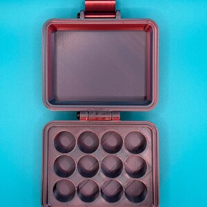 May include: A dark red, rectangular, hinged storage case with a latch. The interior features a flat lid and a base tray with twelve circular, recessed compartments arranged in a three-by-four grid, showing a textured, layered finish.