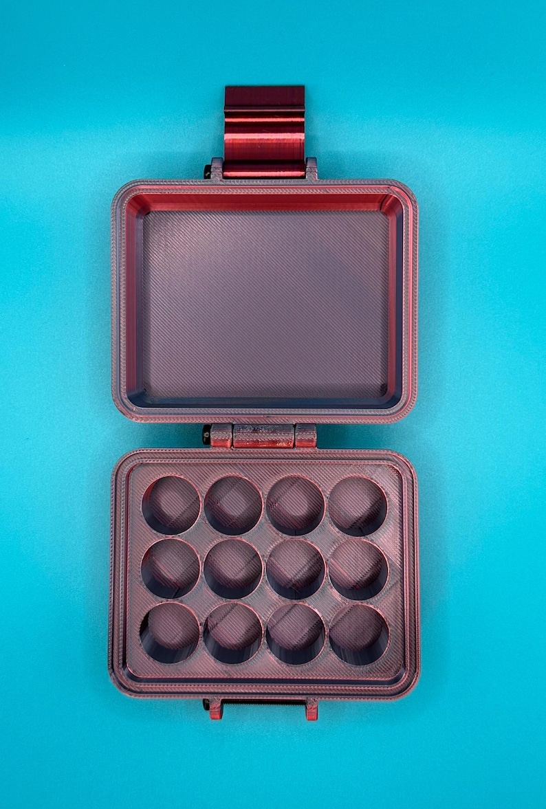 May include: A dark red, rectangular, hinged storage case with a latch. The interior features a flat lid and a base tray with twelve circular, recessed compartments arranged in a three-by-four grid, showing a textured, layered finish.