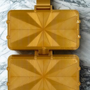 K&ouml;nnte beinhalten: Ein goldfarbenes, rechteckiges, aufklappbares Aufbewahrungsetui mit einem geometrischen Strahlenmuster auf beiden H&auml;lften. Das Etui besteht aus einem starren Material mit sichtbarer Schichtstruktur und einem zentralen Scharnier.