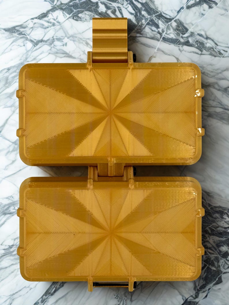 K&ouml;nnte beinhalten: Ein goldfarbenes, rechteckiges, aufklappbares Aufbewahrungsetui mit einem geometrischen Strahlenmuster auf beiden H&auml;lften. Das Etui besteht aus einem starren Material mit sichtbarer Schichtstruktur und einem zentralen Scharnier.