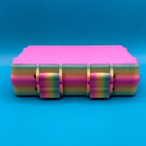 May include: A rectangular storage case featuring a vibrant pink top lid and a multi-colored gradient base that transitions through shades of yellow, green, and blue. The case includes two prominent front latches with matching rainbow colors.