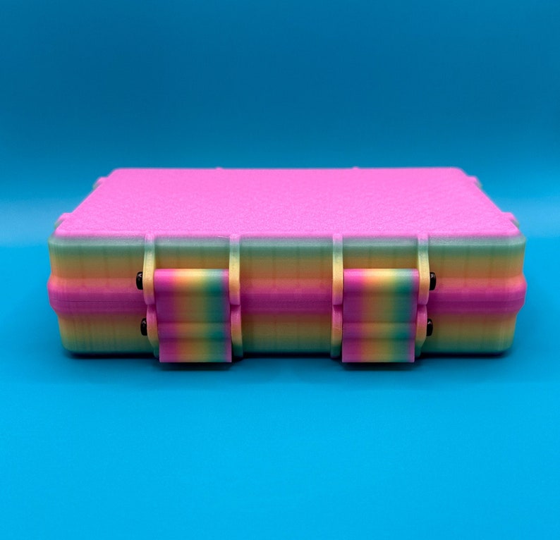 May include: A rectangular storage case featuring a vibrant pink top lid and a multi-colored gradient base that transitions through shades of yellow, green, and blue. The case includes two prominent front latches with matching rainbow colors.