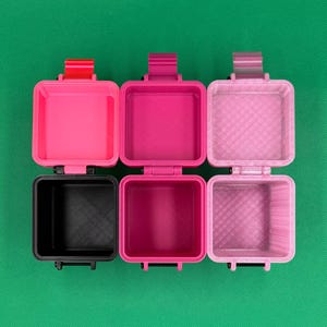 Puede incluir: Tres recipientes cuadrados abiertos con tapas abatibles, dispuestos en fila. El recipiente de la izquierda es negro con una bisagra roja, el del medio es magenta con una bisagra a juego y el de la derecha es de color rosa claro.
