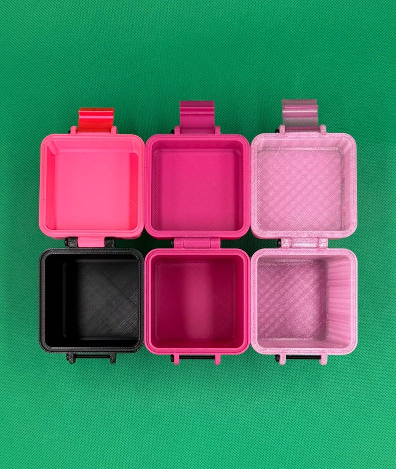 Puede incluir: Tres recipientes cuadrados abiertos con tapas abatibles, dispuestos en fila. El recipiente de la izquierda es negro con una bisagra roja, el del medio es magenta con una bisagra a juego y el de la derecha es de color rosa claro.