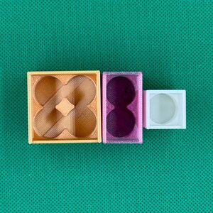 May include: Three 3D printed containers of varying sizes and colors. A large square tan container with a four-lobed interior, a medium rectangular purple container with two circular cutouts, and a small white square container with a circular center.