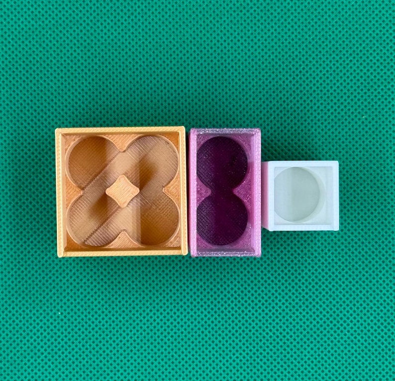 May include: Three 3D printed containers of varying sizes and colors. A large square tan container with a four-lobed interior, a medium rectangular purple container with two circular cutouts, and a small white square container with a circular center.
