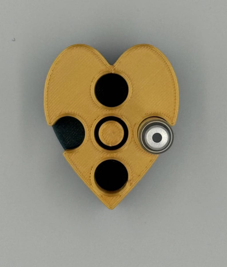 May include: A heart-shaped, mustard-yellow object with three black circular openings and a silver-colored cylindrical component. The object has a textured surface and appears to be 3D-printed.
