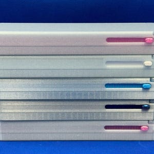 May include: A stack of five rectangular, glittery silver plastic cases. Each case features a horizontal sliding mechanism on the front, with sliders in pink, white, light blue, dark blue, and pink, respectively, set against a blue background.