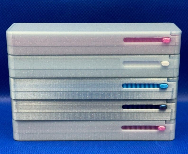 May include: A stack of five rectangular, glittery silver plastic cases. Each case features a horizontal sliding mechanism on the front, with sliders in pink, white, light blue, dark blue, and pink, respectively, set against a blue background.