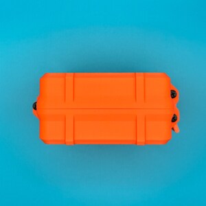May include: A rectangular, bright orange protective storage case featuring a rigid, ribbed exterior design. The case is secured with small black screws visible along the side edge, presenting a durable, utilitarian construction.