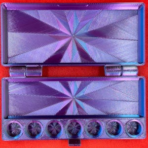 May include: A rectangular, hinged purple and blue iridescent paint palette case with a starburst geometric pattern on the interior lid and base. The bottom section features seven circular wells for paint, all displaying a matching iridescent finish.