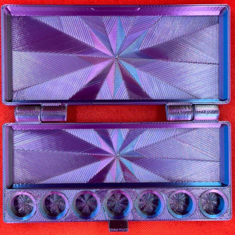 May include: A rectangular, hinged purple and blue iridescent paint palette case with a starburst geometric pattern on the interior lid and base. The bottom section features seven circular wells for paint, all displaying a matching iridescent finish.