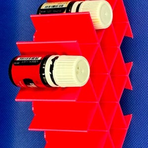 Puede incluir: Un estante de almacenamiento de pl&aacute;stico rojo brillante con una estructura de rejilla de panal para botellas peque&ntilde;as. Dos botellas con tapas blancas y etiquetas negras est&aacute;n insertadas en las ranuras hexagonales. Las etiquetas muestran c&oacute;digos de barras y el texto LOT 134213 y LOT 149567.