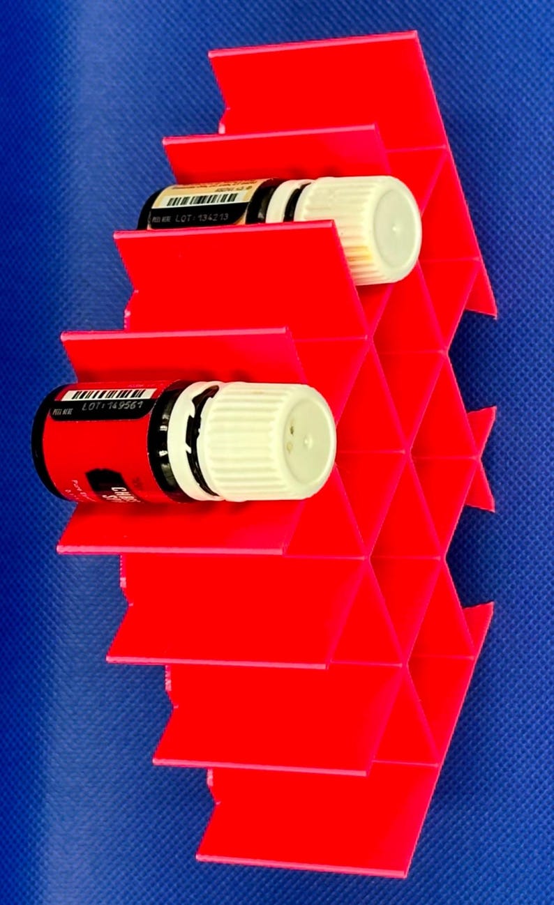Puede incluir: Un estante de almacenamiento de pl&aacute;stico rojo brillante con una estructura de rejilla de panal para botellas peque&ntilde;as. Dos botellas con tapas blancas y etiquetas negras est&aacute;n insertadas en las ranuras hexagonales. Las etiquetas muestran c&oacute;digos de barras y el texto LOT 134213 y LOT 149567.