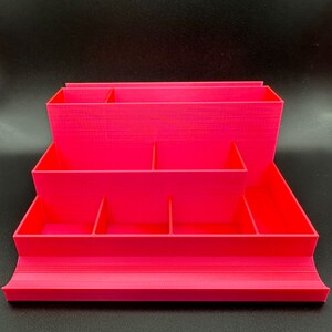 May include: A bright pink tiered desk organizer featuring multiple rectangular compartments of varying heights and depths, including a wide front tray and several vertical dividers for sorting stationery and small office supplies.
