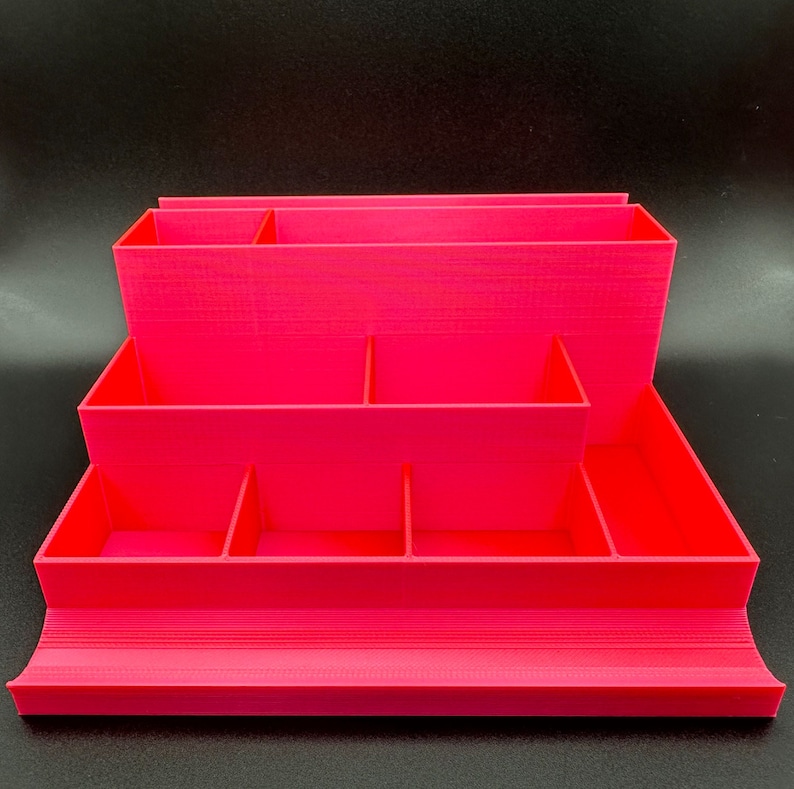 May include: A bright pink tiered desk organizer featuring multiple rectangular compartments of varying heights and depths, including a wide front tray and several vertical dividers for sorting stationery and small office supplies.