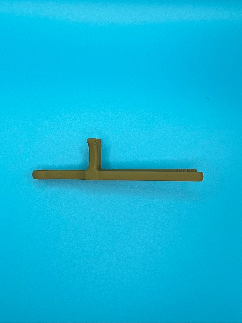 May include: A matte olive green plastic tool featuring a long, flat, horizontal bar with a perpendicular vertical tab extending from the center. The object has a smooth, uniform finish and a simple, functional, linear design structure.