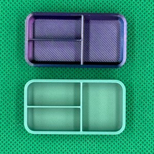 May include: Two rectangular, divided containers. The top container is a gradient of purple and blue, with a textured interior. The bottom container is a solid light green color. Both have rounded corners and are divided into three compartments.