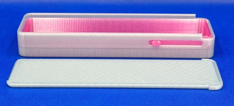 May include: A rectangular, translucent white plastic storage box with a vibrant pink interior lining. The box features a sliding mechanism on the side and comes with a separate, matching translucent white lid that has a textured, cross-hatched surface pattern.