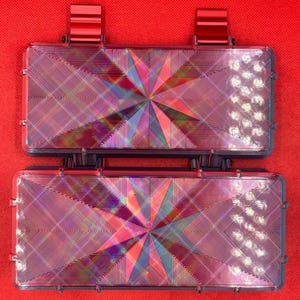 Puede incluir: Dos cajas de almacenamiento rectangulares de color oscuro con tapas abatibles, cada una con una superficie superior hologr&aacute;fica con patr&oacute;n de estrella que refleja la luz en varios colores. Tienen clips met&aacute;licos rojos en los bordes.